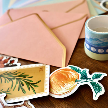 Sticker with a orange fruit design on a wooden surface with envelopes and a stack of washi tape rolls.